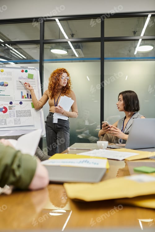 Preview: redhead businesswoman pointing at infographics on flip chart near colleagues in meeting room