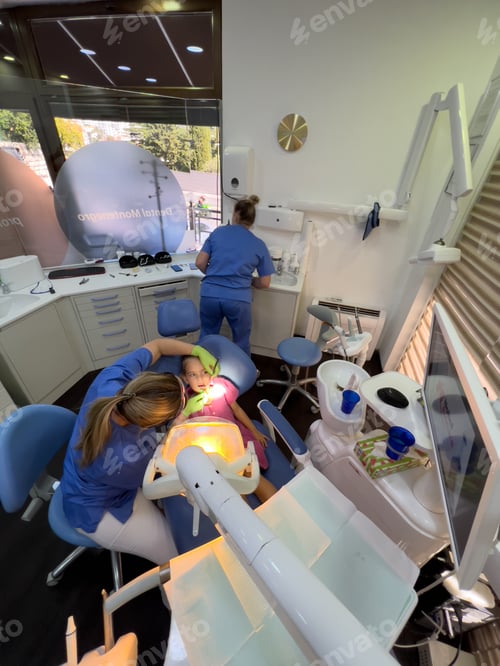 Preview: Dentist examines with a mirror under the lamp a little girl sitting with her mouth open