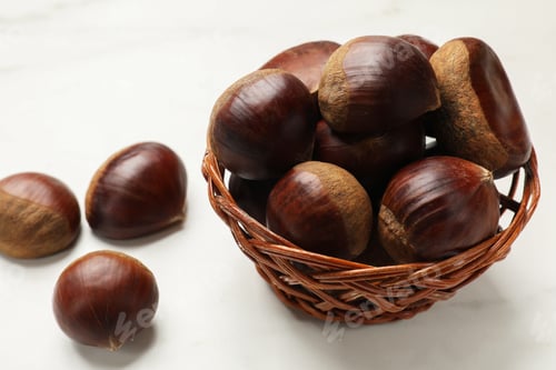 Preview: Sweet fresh edible chestnuts in wicker bowl on white table, closeup