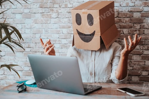 Preview: Happy modern online worker wearing smile carton box on head speaking in video call at the laptop