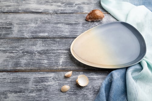 Preview: Empty blue ceramic plate on gray wooden background and blue textile. Side view, copy space.