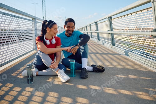 Preview: Smiling athletic couple using smart watch while resting after sports training outdoors.