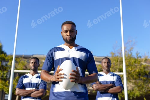 Preview: Three African American young male athletes holding a rugby ball, standing on field outdoors