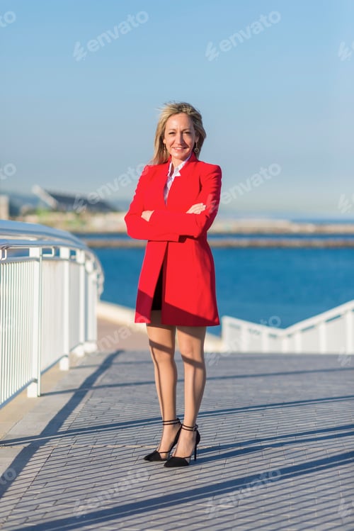 Preview: Adult woman standing on a bridge next to the sea while crossing arms and looking directly to camera
