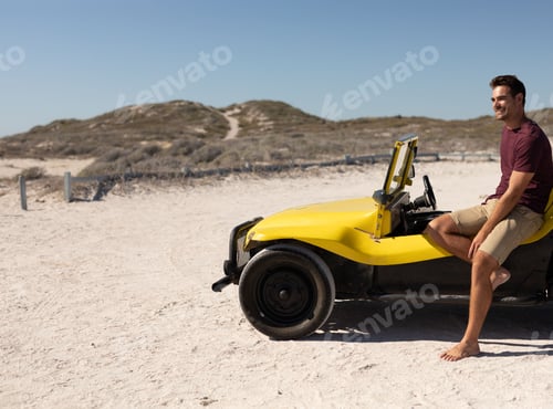 Preview: Man sitting barefoot on yellow dune buggy in coastal dunes with wooden posts, copy space