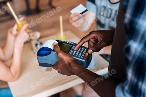 Preview: cropped view of african american waiter with terminal taking payment with credit card in cafe
