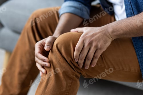 Preview: Knee pain, close-up of a man massaging the knee joint of his leg while sitting on a sofa in the