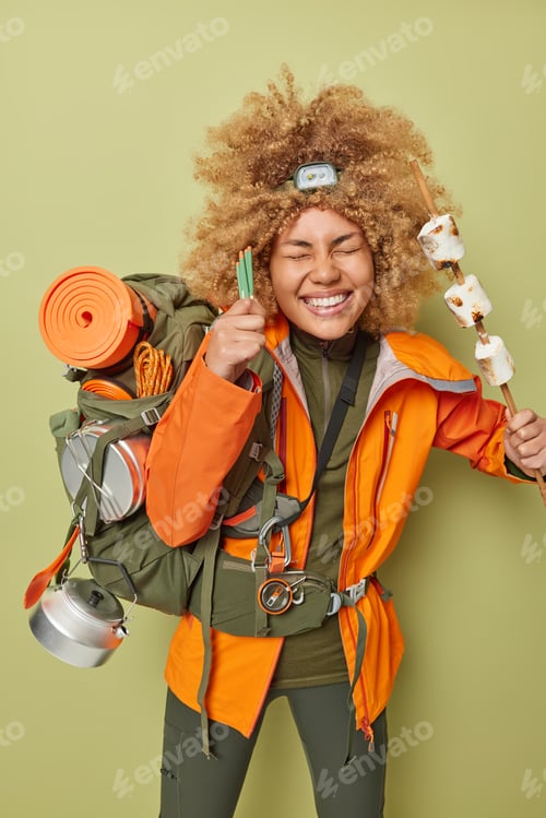 Preview: Cheerful female camper holds roasted marshmallow has picnic during spare time wears orange jacket re