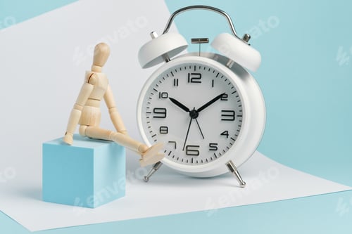 Preview: Wooden man sits near a white alarm clock. Concept of watching time, long wait.