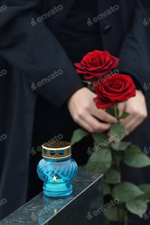 Preview: Woman holding red roses near black granite tombstone with candle outdoors, closeup. Funeral ceremony