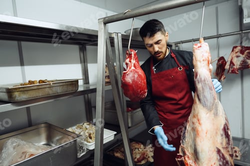 Preview: Male butcher standing by meat hanging on hook in meat shop