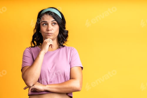 Preview: Pensive young woman reflecting with hand on chin on yellow background