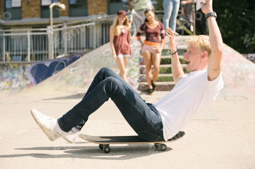 Preview: Young male skateboarder sitting on skateboard on the move in city skatepark