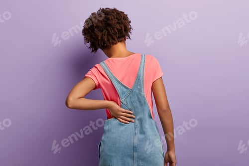 Preview: Young woman with curly hair, suffers from radiculitis, keeps hand on waist, wears pink t shirt and d