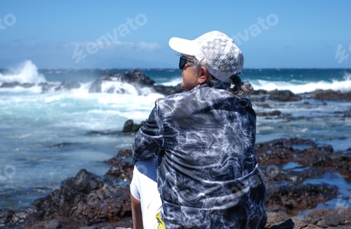 Preview: Rear view of Mature woman sitting on a rocky beach while admiring the sea waves