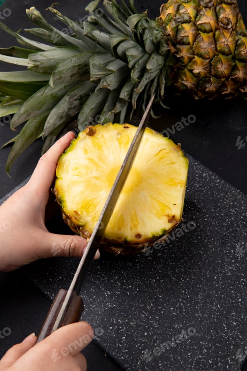 Preview: side view of woman hand cutting pineapple with knife on cutting board on black background