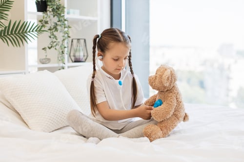 Preview: Girl using stethoscope to check toy bear on bed