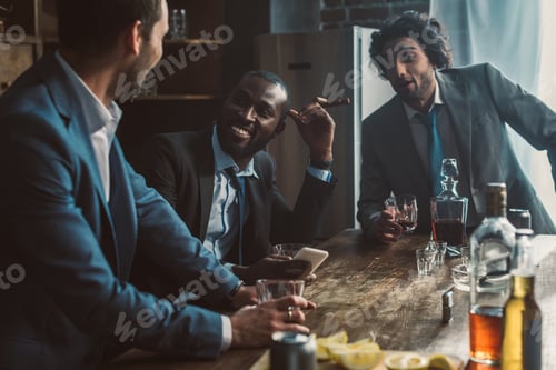 Preview: smiling multiethnic men talking, drinking whiskey and smoking cigars while partying together