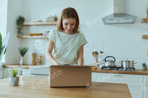 Preview: Attractive young woman unpacking box while standing at the domestic kitchen