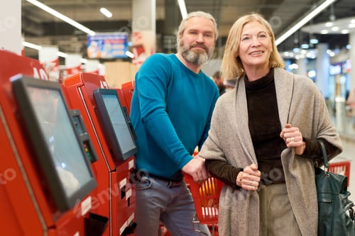 Preview: Happy aged couple in smart casualwear standing by self service terminals