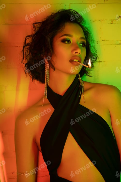 Preview: Sexy asian woman in dress holding match with fire in lips while standing in night club with lighting