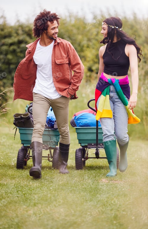 Preview: Young Couple Pulling Trolley Filled With Camping Equipment Through Field To Music Festival