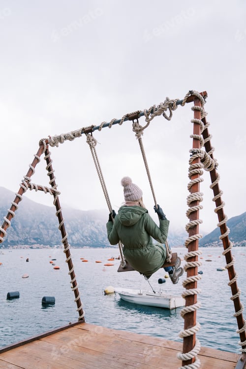 Preview: Young woman swings on a swing on the pier and looks at the mountains. Side view