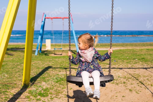 Preview: Adorable baby girl wearing a white dress enjoying a swing ride on a playground in a park on a nice