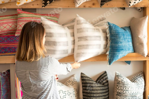 Preview: Rear view of female interior designer adjusting cushions on shelves in retail studio