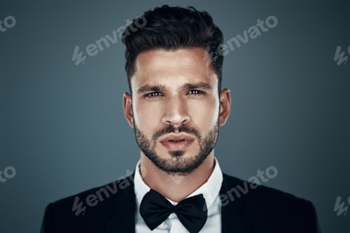 Preview: Charming young man in bow tie looking at camera while standing against grey background