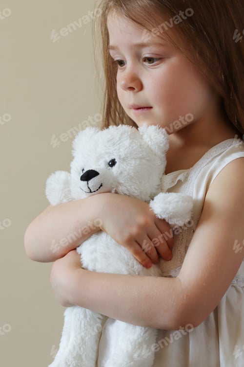 Preview: A little girl in a white dress holds a teddy bear in her hands