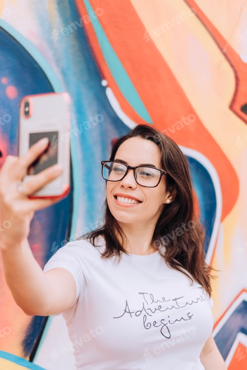 Preview: Happy woman smiling wearing glasses and taking selfie on mobile phone in front of colorful wall art