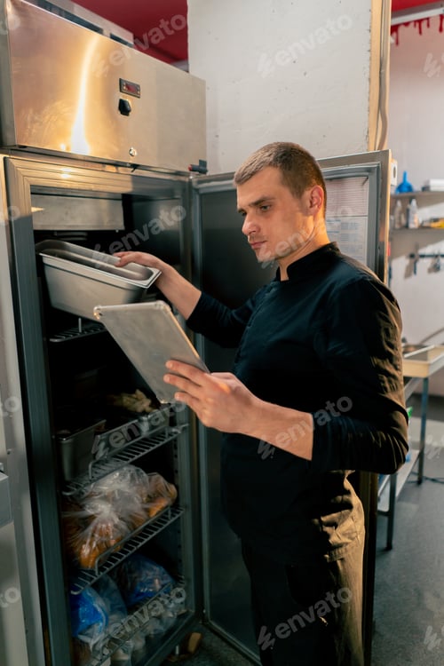Preview: chef stands in kitchen in a black uniform and looks through what is on the list in the refrigerator