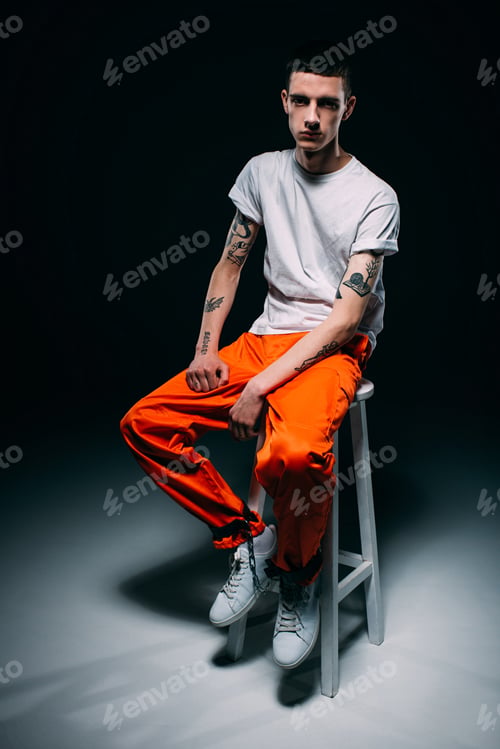 Preview: Serious man in orange uniform with cuffs on legs sitting on stool on dark background
