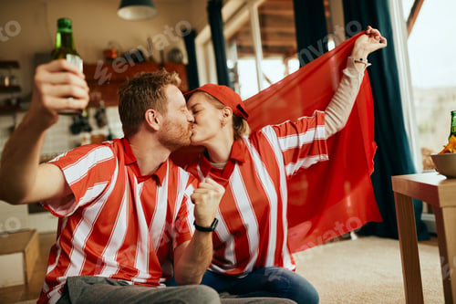 Preview: Happy couple kissing while watching sports match on TV and celebrating victory of their team.