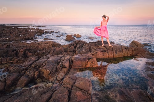 Preview: Beautiful young girl in pink dress standing on the rock with reflecions in the water at Wild atlanti