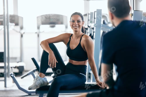 Preview: A smiling girl sitting on a bench and talking to her gym partner, resting together.