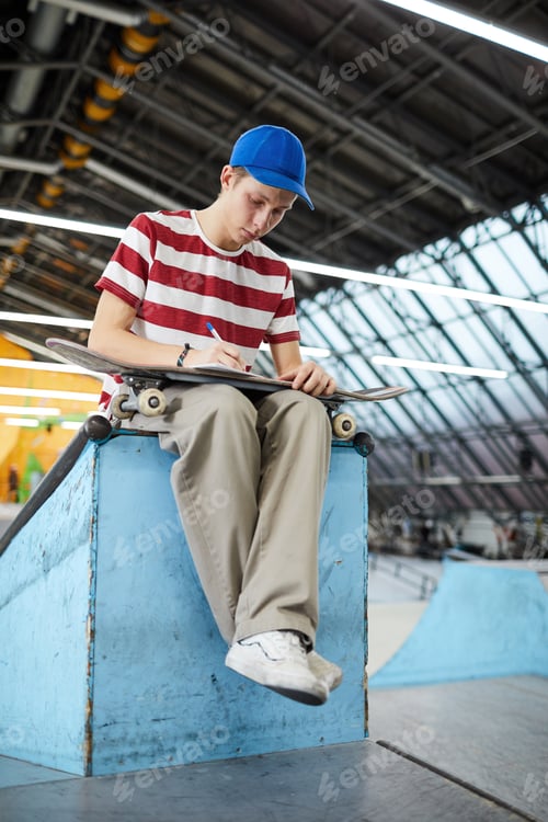 Teenager Sits at Skate Park Writing on Skateboard