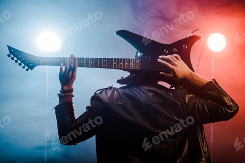 Preview: rear view of male musician in leather jacket playing on electric guitar behind head on stage