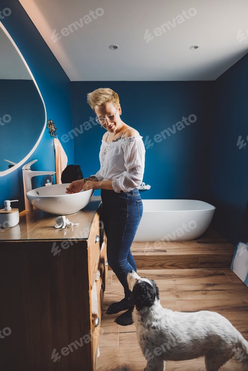 Visualização: Uma mulher com cabelo loiro curto está em um banheiro moderno, sorrindo enquanto lava as mãos. Um preto