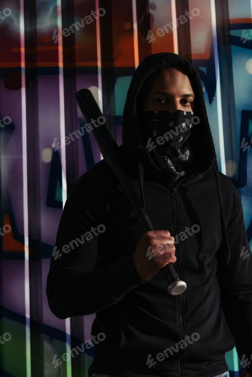 Preview: African american hooligan holding baseball bat and looking at camera near graffiti on urban street