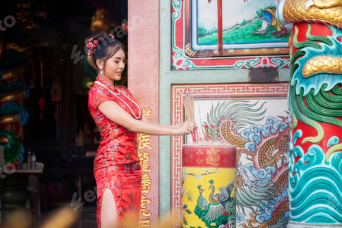 Preview: Chinese woman in a red cheongsam dress holding incense pay homage to Chinese god at shrine.
