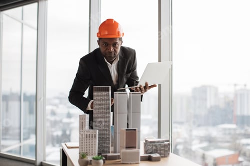 Preview: Focused african inspector using laptop inspecting skyscraper model in architectural modern office