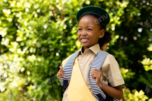 Preview: Smiling african american scout girl in uniform with backpack looking away against plants