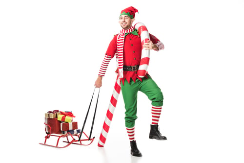 Preview: happy man in christmas elf costume carrying sleigh with presents and big candy cane isolated on