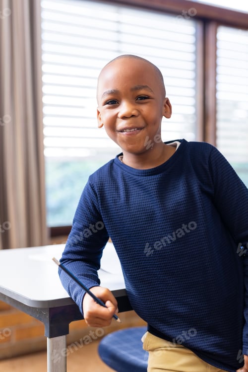 Preview: Smiling african american boy holding pencil and leaning on desk in school classroom