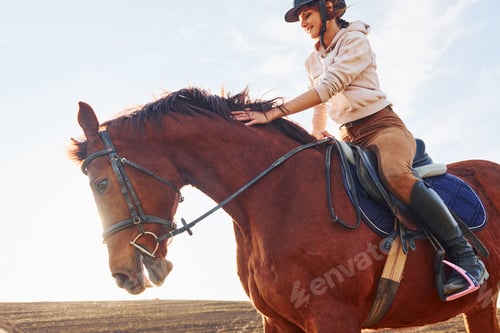 Preview: Young woman in protective hat with her horse in agriculture field at sunny daytime