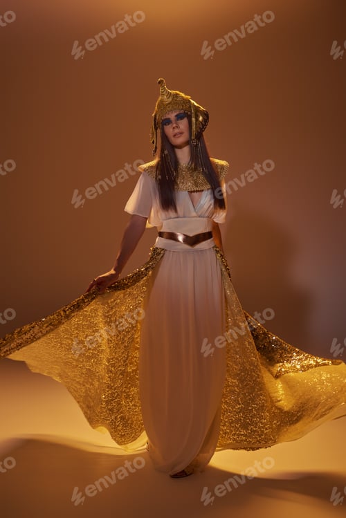Preview: Full length of woman in egyptian attire holding elegant skirt while standing on brown background