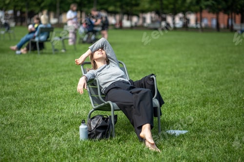Preview: Work-weary woman take break relaxed reclines on chair in open public space on lawn hands over eyes.