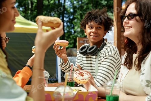 Preview: Smiling Teenage Black Boy Holding Donut at Table with Friends Outdoors